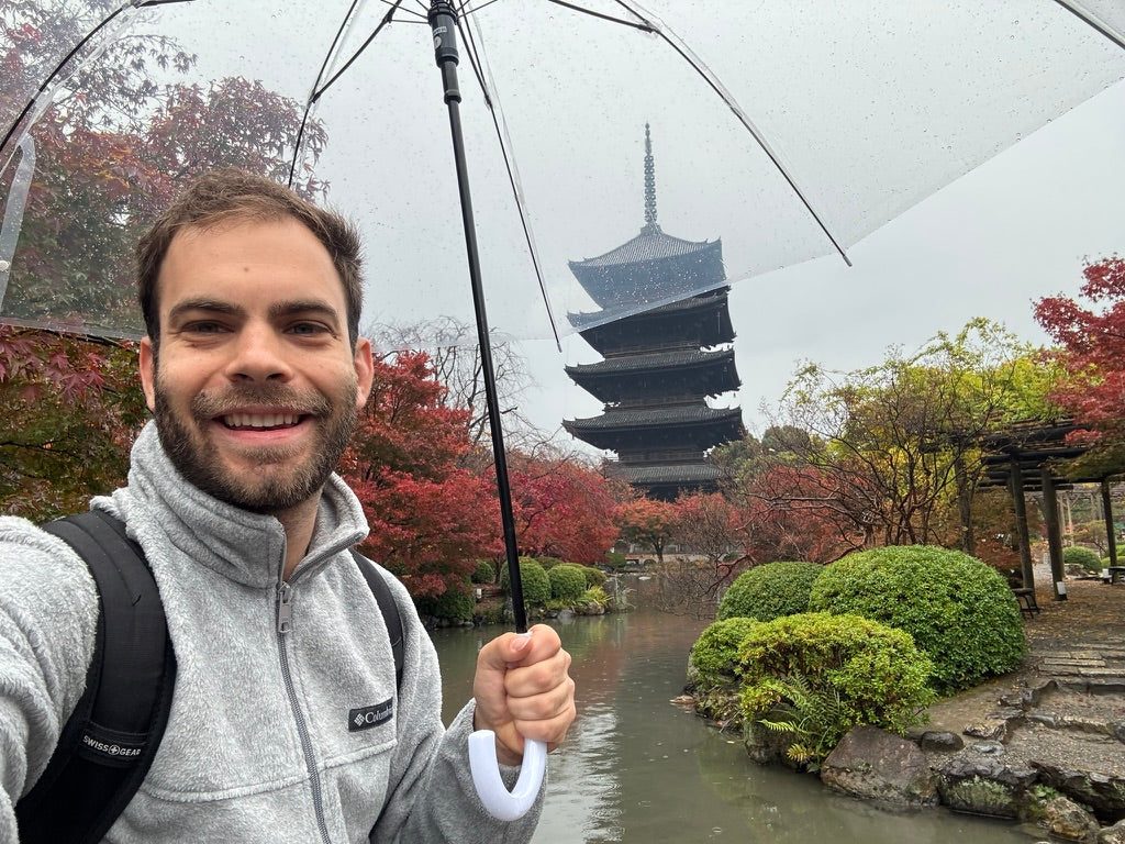 A close-up selfie of a smiling man with a beard, wearing a grey fleece jacket and holding a transparent umbrella, with a view of the five-story Toji Temple pagoda and vibrant red and green autumn trees in Kyoto, Japan, in the background.