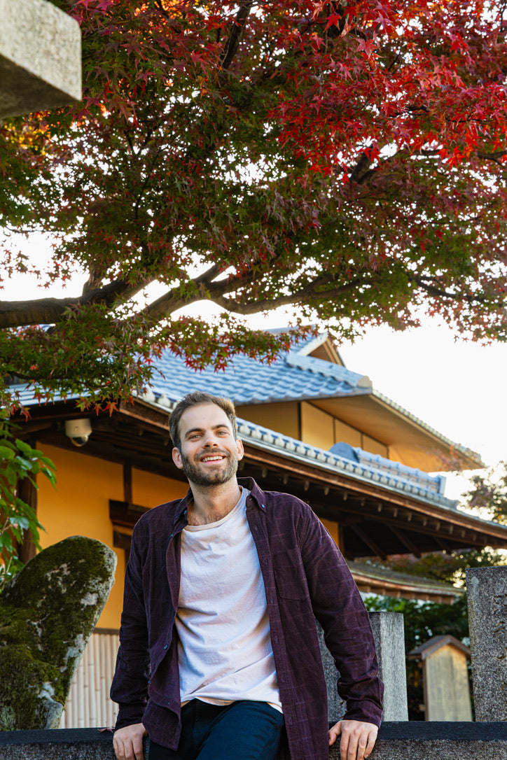 A portrait of a man smiling in front of a traditional Japanese building with a blue-tiled roof, framed by vibrant red autumn maple leaves under a clear sky.